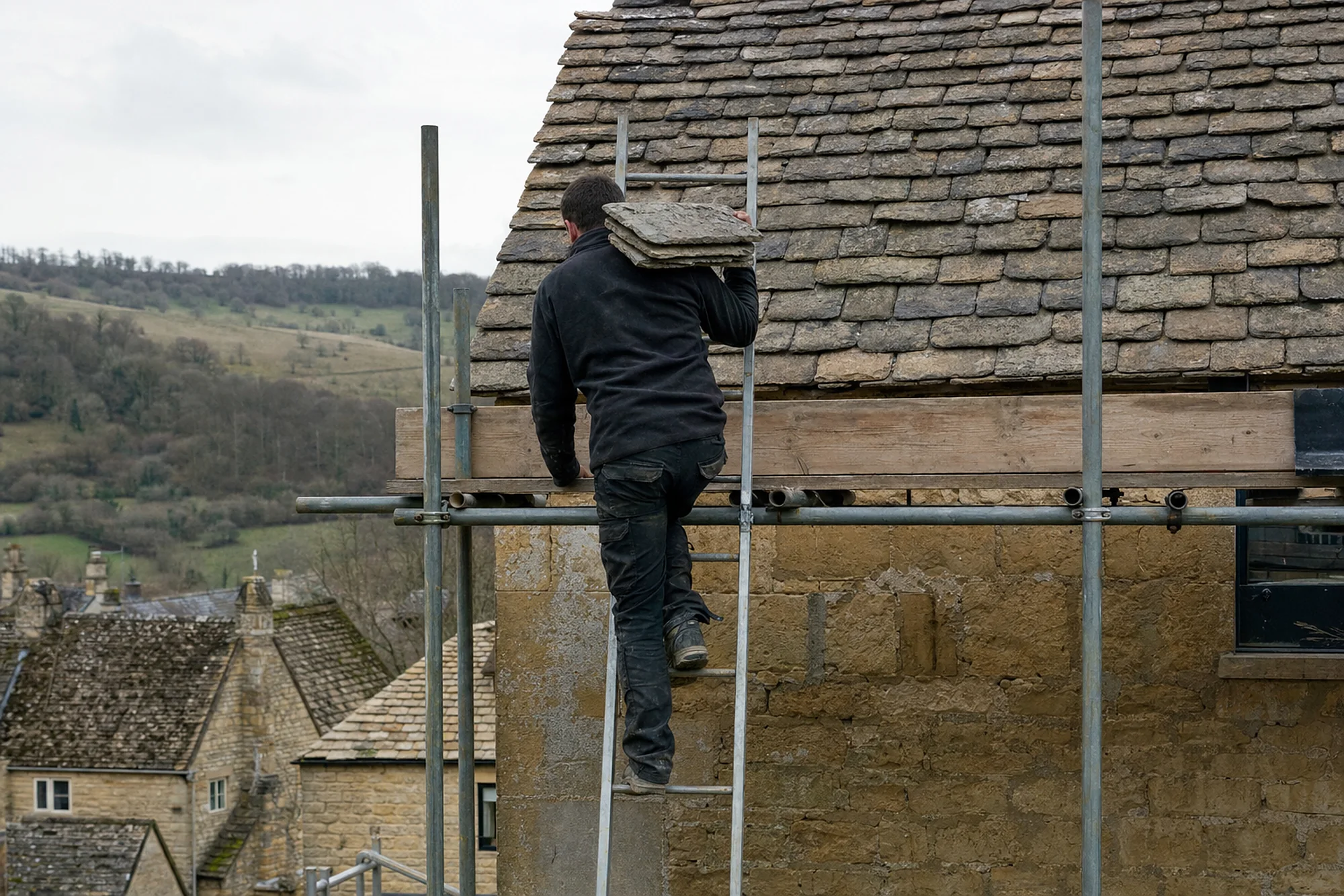 Roofer climbing a scaffold ladder with Cotswold stone slates on his shoulder beside a stone slate roof