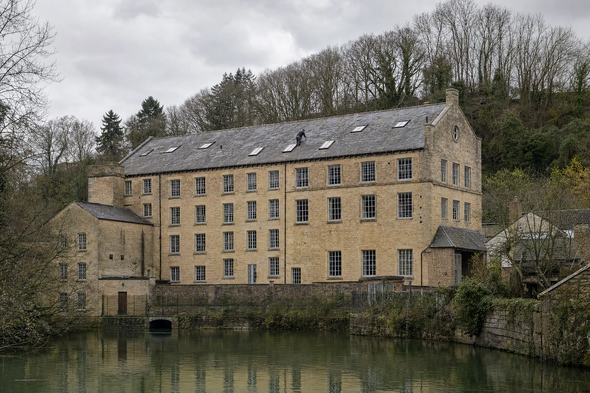 Large stone woollen mill with a long slate roof and rooflights beside still water in a wooded valley