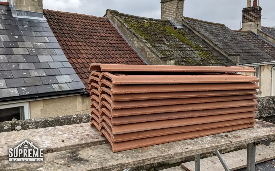 Stack of red clay ridge tiles on a scaffold board beside slate roofs