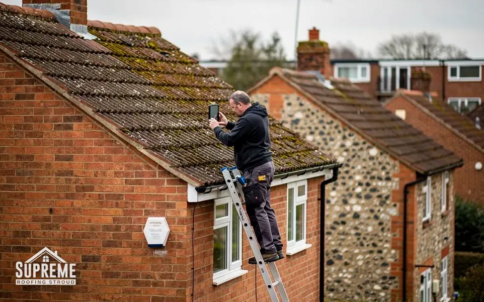 Man on a ladder photographing a mossy tiled roof on a brick house