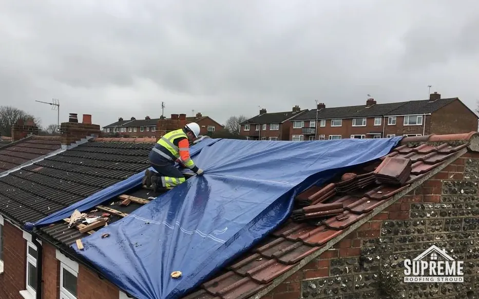 Roofer securing a blue tarpaulin over damaged roof tiles on a terrace