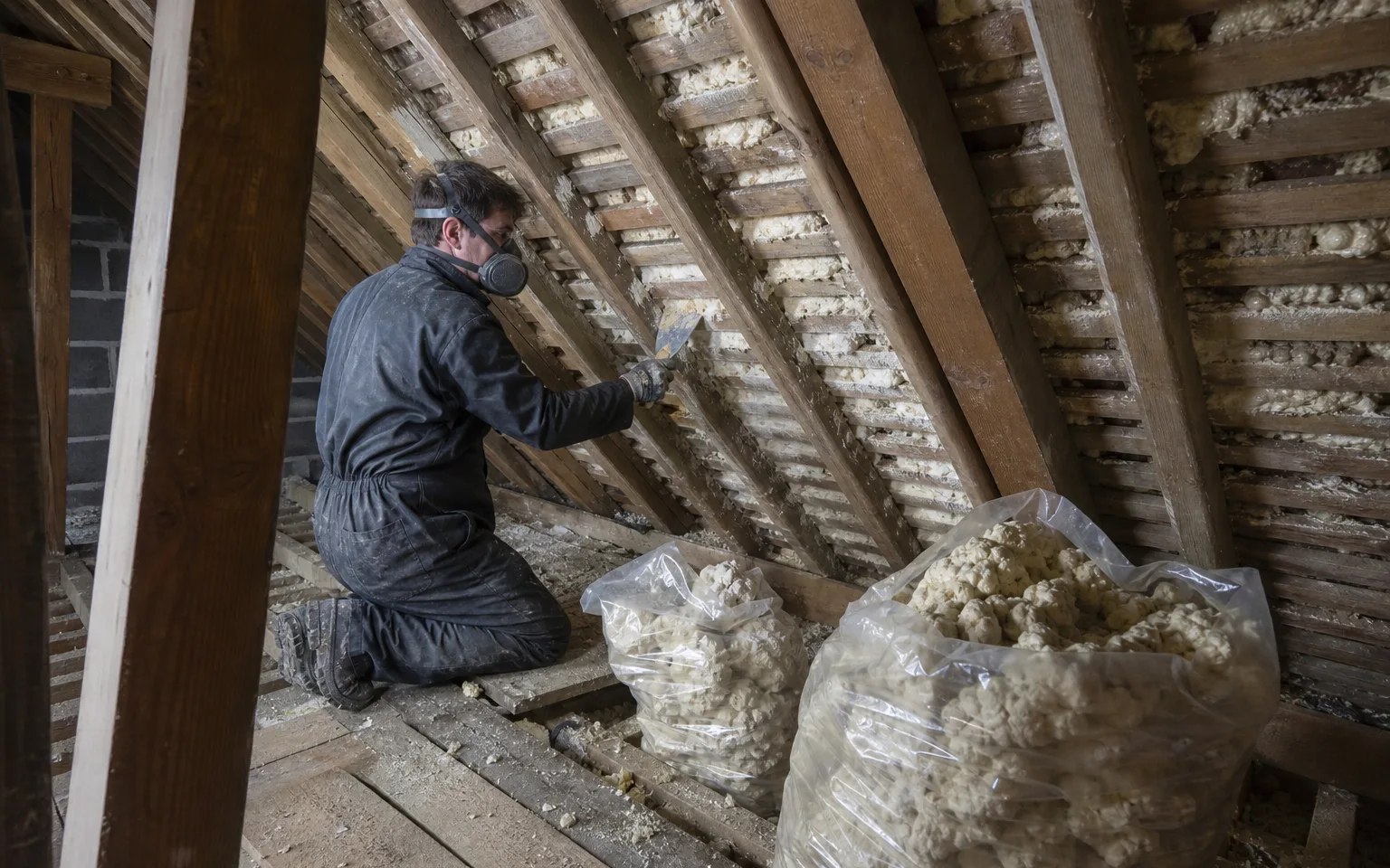Worker in a mask scraping spray foam insulation off roof timbers in a loft